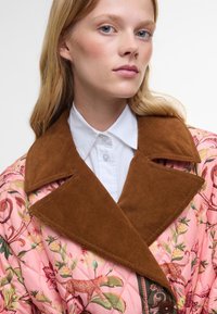 Prošívaná růžová bunda s květinovými a zvířecími vzory, se hnědým kordovým límcem a knoflíkovou detailou, pod kterou je bílé tričko.