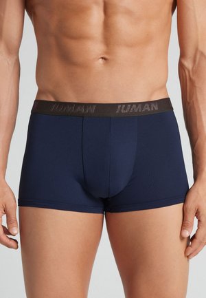 Mandlig torso iført tætsiddende navyblå boxer briefs med en sort taljebånd med subtil tekst på en neutral baggrund.