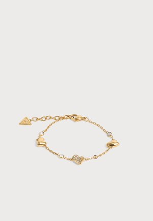 Bracelet chaîne en or avec trois charms en forme de cœur, dont un recouvert de cristaux, et trois petites pierres rondes transparentes, sur un fond clair.