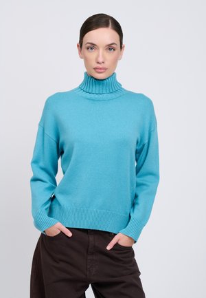 Mujer de piel clara y ojos azules que lleva un jersey de cuello alto azul y pantalones oscuros, está de pie con las manos en los bolsillos frente a un fondo liso.