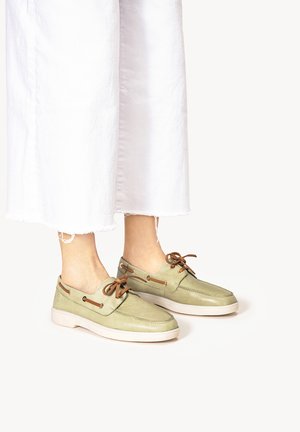 Chaussures bateau en daim vert avec lacets marron et accents en métal, dotées d'une semelle en caoutchouc de couleur claire et d'un bout arrondi. Portées avec un pantalon blanc coupé au-dessus de la cheville.