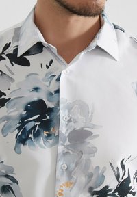 Chemise blanche avec un motif floral dans des nuances de bleu et de gris. Le tissu est lisse avec un devant à boutons et un col pointu.
