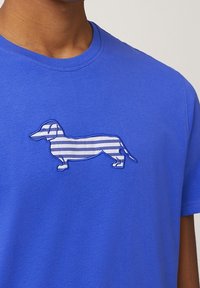 Blå bomulls t-shirt med en broderad kontur av en tax med horisontella vita ränder inuti konturen.