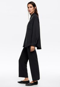 Camisa negra de tejido entrelazado con botones al frente y bolsillo en el pecho, combinada con pantalones negros de corte relajado y zapatos planos negros. Textura suave, diseño minimalista.