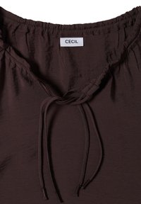 Blouse marron foncé avec encolure élastique et lien devant, avec une étiquette blanche portant le nom de la marque "CECIL" cousue à l'intérieur du col.