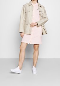 Light beige jacket with front pockets over a light pink button-up dress, paired with white sneakers. Smooth textures, contemporary design.