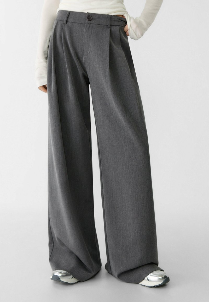 Hüftlange Hose aus einem dunkelgrauen Stoff, mit gefalteter Vorderseite, einem einzelnen Knopfverschluss und einer glatten Textur.