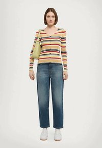 Naine kannab triibulist nööbiga pikkade käistega särki, siniseid teksaseid, valgeid spordi jalanõusid ja tal on õlgadel kerge rohelise värvi kott, seismas ühtlase tausta vastu.