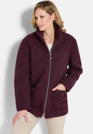 Veste en polaire bordeaux avec col montant, fermeture éclair et deux poches avant. Texture douce, coupe décontractée et manches longues avec bords côtelés.