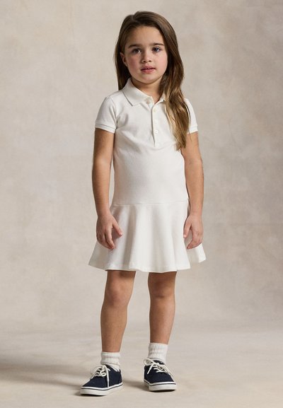 Young girl standing, wearing a white short-sleeve polo dress, white socks, and dark blue sneakers against a neutral background.