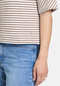 Kortærmet top med et hvidt og brunt diamantmønster, parret med blå denim jeans. Toppen har en diskret logo-detalje.