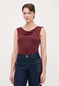 Top senza maniche color bordeaux con scollatura drappeggiata, abbinato a jeans in denim scuro a vita alta con dettagli in pizzo sui lati.