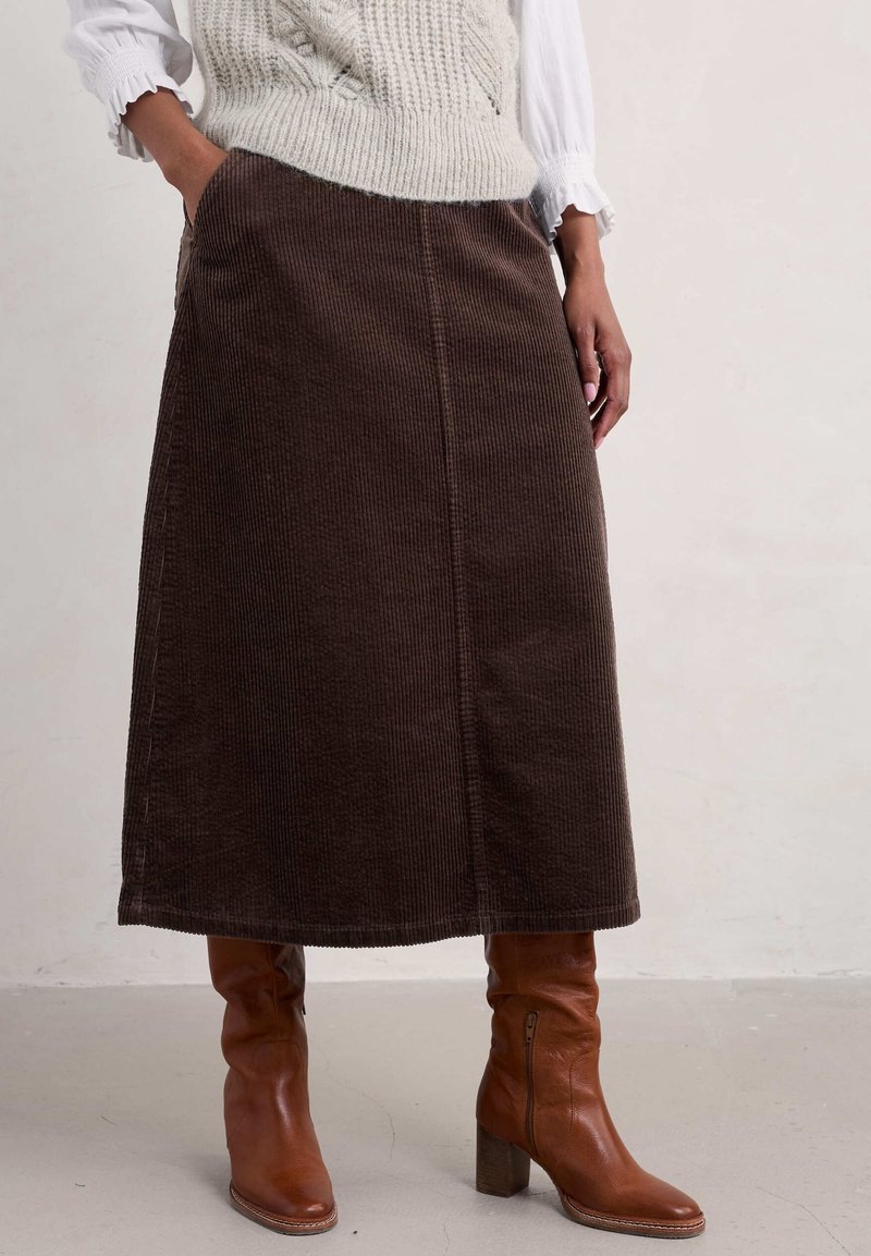 Femme portant une jupe midi en velours côtelé marron avec des poches, associée à des bottes à talons en cuir marron et un gilet en maille crème porté par-dessus une chemise blanche.