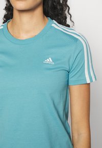 Ljusblå bomullströja med rund halsringning, korta ärmar och vita tre-strimlor på axlarna. Litet vitt Adidas-logotyp på bröstet.