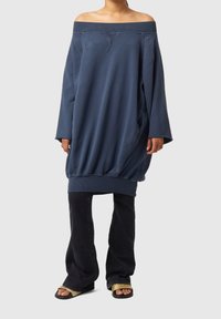 Översized off-shoulder sweatshirt i marinblått, med en ribbad halsringning och texturerad fåll, kombinerad med mörka utsvängda jeans och enkla sandaler.