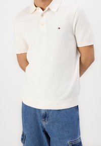 Muž v bílém polo tričku s krátkým rukávem a malým logem na hrudi, v modrých džínových cargo šortkách, s rukama za zády.