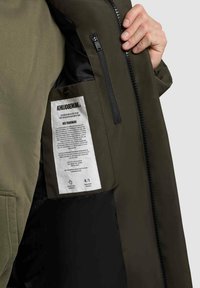 Unutrašnjost maslinasto zelene jakne s crnim podlogom, koja ima etiketu, zip džep i čvrsto zatvaranje sa patentnim zatvaračem. Vidljiva teksturirana tkanina.