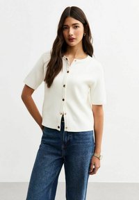 Cardigan bianco a maniche corte con scollatura rotonda e chiusura a bottoni, dotato di bottoni tono oro, abbinato a jeans blu a vita alta.