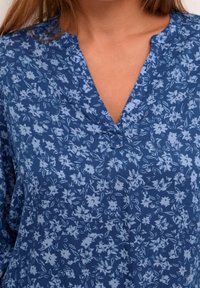 Blaue Bluse mit floralem Muster, V-Ausschnitt und lockerer Passform. Der Stoff zeigt kleine, hellblaue Blumenmuster auf einem dunkleren blauen Hintergrund.