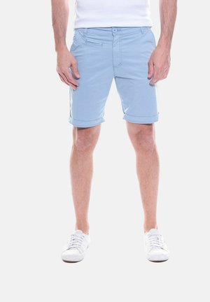 Homme portant un short bleu clair retroussé, des baskets blanches et une chemise blanche, debout devant un fond uni.