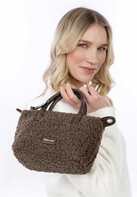 Mujer rubia con un suéter blanco sosteniendo un pequeño bolso marrón texturizado con asas de cuero sobre su hombro.