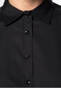 Gros plan sur une chemise noire à boutons avec un col pointu et quatre boutons noirs visibles le long de la patte de boutonnage.
