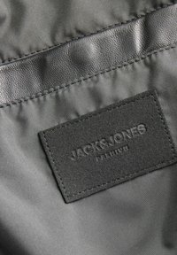 Crni materijal s glatkom teksturom sadrži ušivenu tamnosivu oznaku brenda "JACK & JONES PREMIUM" sa utisnutim slovima.