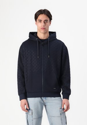 Marineblauwe zip-up hoodie met een gestructureerd geometrisch patroon op het bovenste gedeelte, voorzien van een kangoeroezak en een trekkoord capuchon.