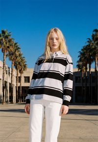 Femme blonde debout à l'extérieur portant une chemise à manches longues rayée noir et blanc et un pantalon blanc, avec des palmiers et des bâtiments en arrière-plan.