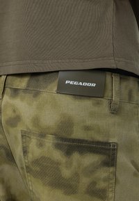 Pantalones de camuflaje verde con una etiqueta de cuero PEGADOR en la cintura. La tela parece texturizada con patrones sutiles y detalles de costura.