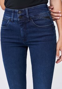 Vaqueros de mezclilla oscuros de tiro alto, con dos botones negros, bolsillos delanteros y una textura suave. Detalles de costura a lo largo de las costuras.