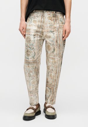 Mann trägt gemusterte Hose mit Sonnen- und Blumenmotiven, kombiniert mit beige-braunen Loafers mit goldenen Ketten.