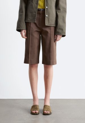 Persona con chaqueta oliva, camisa amarilla, pantalones cortos marrones hasta la rodilla con costuras delanteras y zapatos oliva abiertos sin cordones, de pie sobre suelo gris.