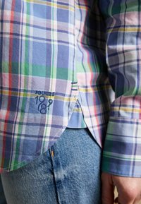 Tuva cilvēka tuvplāns, kurš valkā krāsainu pleda raksta kreklu ar izšūtu uzrakstu "Joules 1989" pie apakšmalas un zilos džinsa bikses.