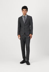 Costume gris foncé avec un tissu texturé, veste à un bouton, et pantalon ajusté. Associé à une cravate grise claire et des chaussures noires.