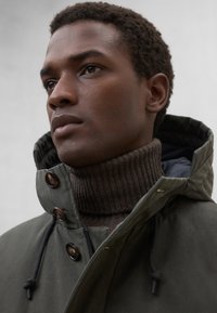 Oliwkowa parka z wysokim kołnierzem, pod którym znajduje się brązowy prążkowany golf. Posiada ciemne guziki i sznurowane detale.