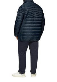 Navy Pufferjacke mit einem gesteppten Design, hohem Kragen und dezentem Branding; kombiniert mit dunklen Hosen und weißen Sneakers.
