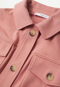 Gros plan sur une chemise en tissu rose texturé avec un col, deux poches avant, et de gros boutons beiges avec des accents foncés.
