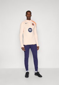 Long-sleeve peach athletic top with a half-zip, featuring a Spotify logo and FC Barcelona crest, paired with navy sweatpants and black shoes.