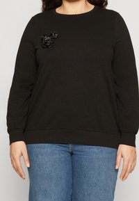 Black sweatshirt made of soft material, featuring a round neckline and long sleeves. Three black fabric flowers adorn the chest area.