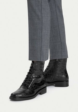 Czarne, błyszczące sznurowane botki w wzór krokodyla z bocznym zamkiem, noszone z szarymi, dopasowanymi spodniami.