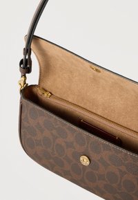 Καφέ τσάντα ώμου με σχέδιο, κούμπωμα με χρυσό κουμπί, εσωτερικό από ταμπά σουέτ και εσωτερική τσέπη με φερμουάρ μερικώς ανοιχτή σε λευκό φόντο.