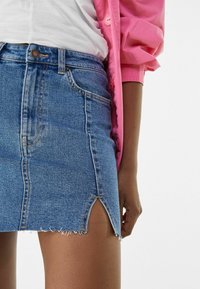 Džínová sukně v středně modré barvě s třepícím se lemem a bočními rozparky. Obsahuje pět kapes a hnědý prošívání na švech. Nosená s bílým topem.