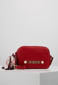 Punainen keinonahkainen olkalaukku, jossa on kultainen logo ja hopeiset metalliosat sekä irrotettava kuvioitu hihna, jossa on huulipunateemaisia kuvioita.