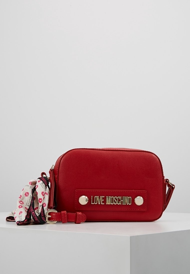 Punainen keinonahkainen olkalaukku, jossa on kultainen logo ja hopeiset metalliosat sekä irrotettava kuvioitu hihna, jossa on huulipunateemaisia kuvioita.