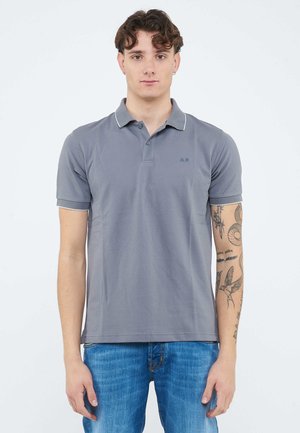 Giovane uomo con capelli corti e ricci che indossa una polo grigia e jeans blu, mostrando i tatuaggi sull'avambraccio destro.