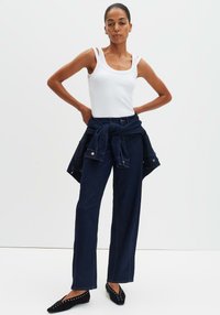 Canotta bianca con spalline sottili, abbinata a jeans blu navy a gamba larga e giacca di jeans annodata in vita. L'outfit è completato da ballerine nere con borchie.