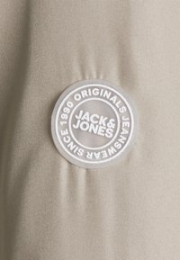 Beżowy materiał o gładkiej fakturze z okrągłą plastikową naszywką. Naszywka zawiera biały napis: "JACK & JONES ORIGINALS JEANSWEAR OD 1990 R."
