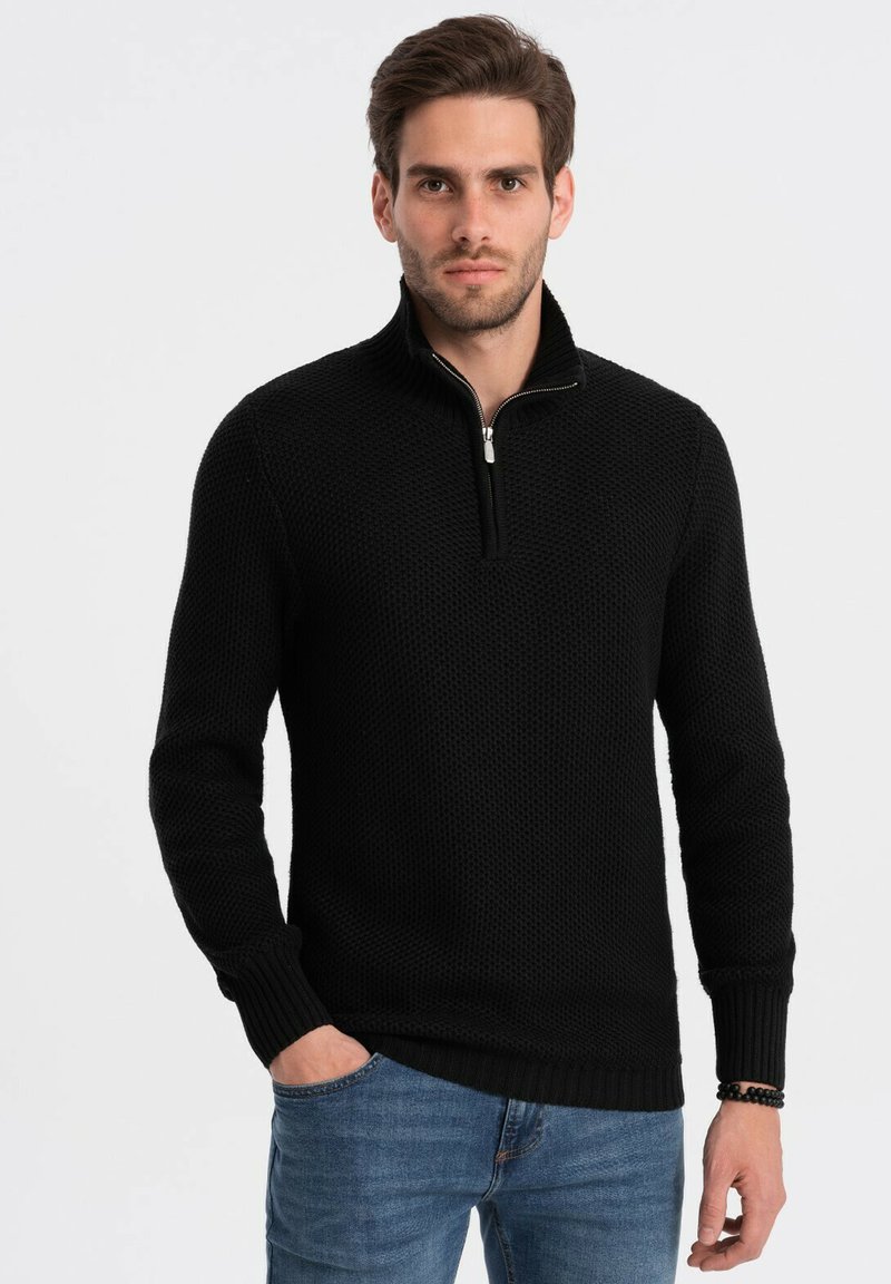 Maglione nero lavorato a maglia con collo alto e zip frontale a metà. Presenta polsini e orlo a costine. Indossato sopra un paio di jeans blu, con una finitura testurizzata.