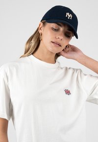 Mujer que lleva una camiseta blanca con un pequeño logo rojo y azul y una gorra de béisbol azul marino con letras bordadas blancas "NY".
